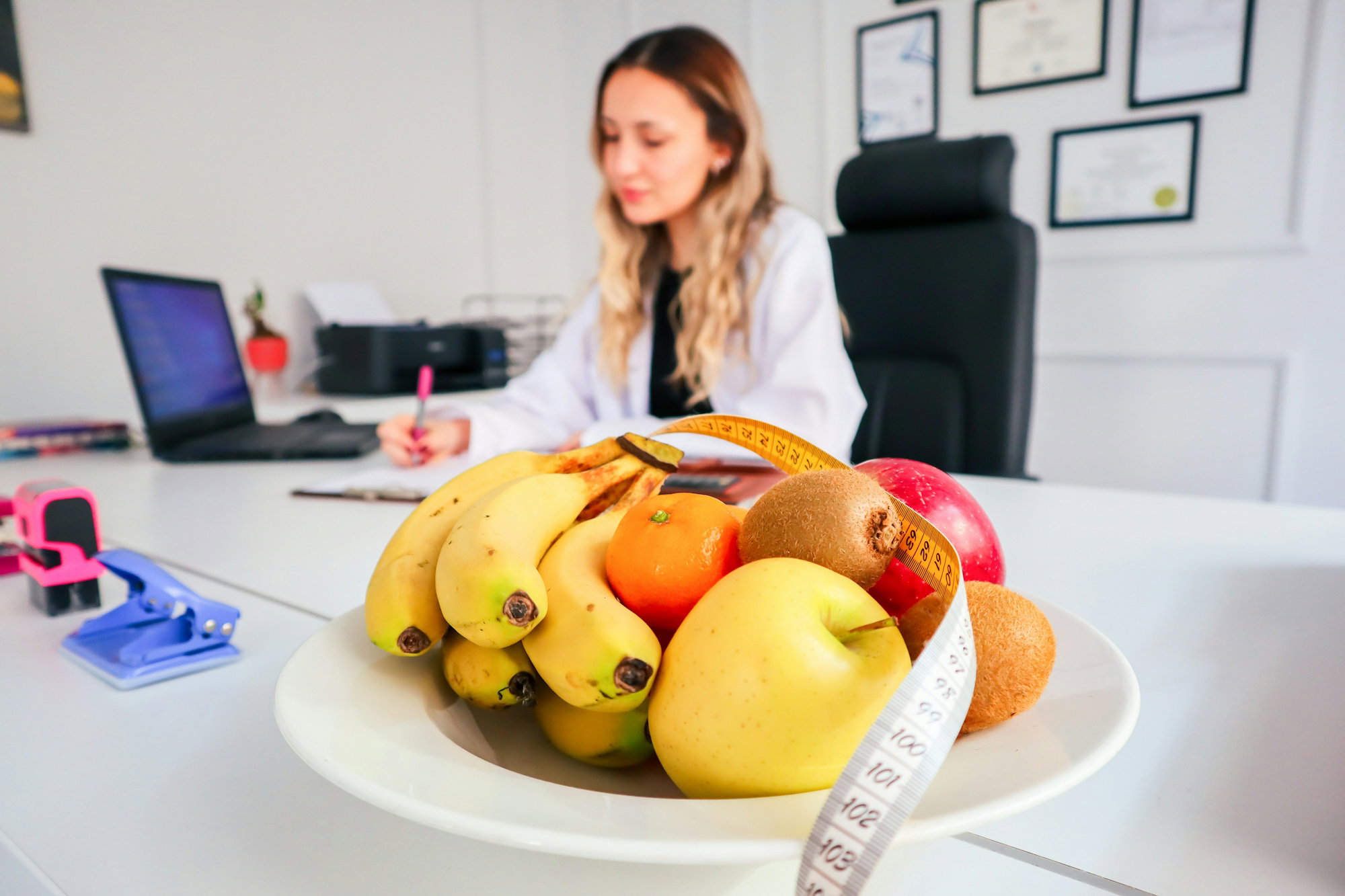 a-woman-sitting-at-a-desk-with-a-bowl-of-fruit.jpg Nutricionista em consultório com frutas sobre a mesa